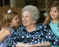 Elderly woman in wheelchair receiving kisses from her granddaughters at Manoogian Manor.