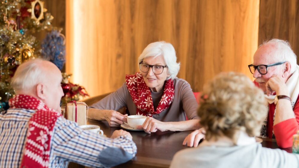 Group of positive elderly friends in scarves with Christmas patterns sitting at table and chatting while drinking coffee at Christmas eve