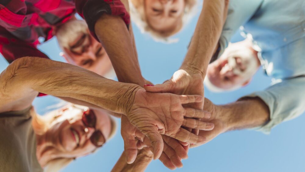 Low angle view of group of cheerful active senior people enjoying sunny summer day outdoors, gathered in circle, holding hands all together in the middle