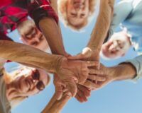 Low angle view of group of cheerful active senior people enjoying sunny summer day outdoors, gathered in circle, holding hands all together in the middle