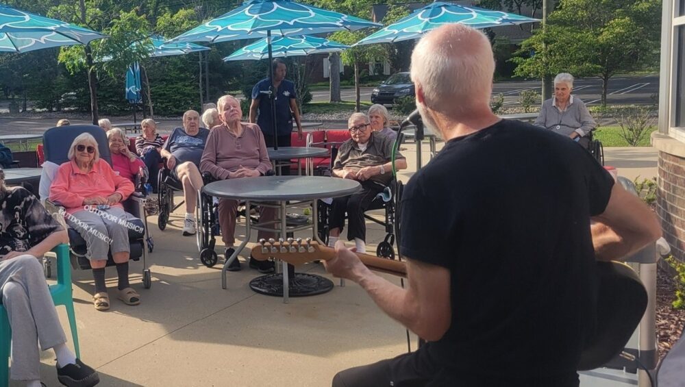 Photo of a man playing a guitar for a group of senior citizens on the Manoogian Manor patio.