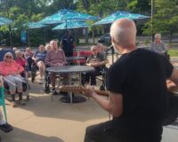 Photo of a man playing a guitar for a group of senior citizens on the Manoogian Manor patio.