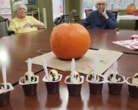 Close-up of Halloween desserts with a pumpkin in the middle of the table and senior citizens in the background.