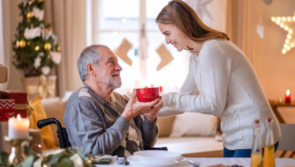 Granddaughter giving hot cocoa to grandfather in wheelchair.