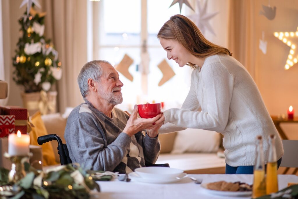 Granddaughter giving hot cocoa to grandfather in wheelchair.