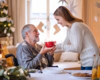 Granddaughter giving hot cocoa to grandfather in wheelchair.