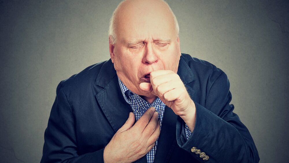 Old man coughing holding fist to mouth isolated on gray background