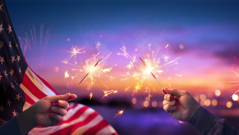USA Celebration With Hands Holding Sparklers And American Flag At Sunset With Fireworks
