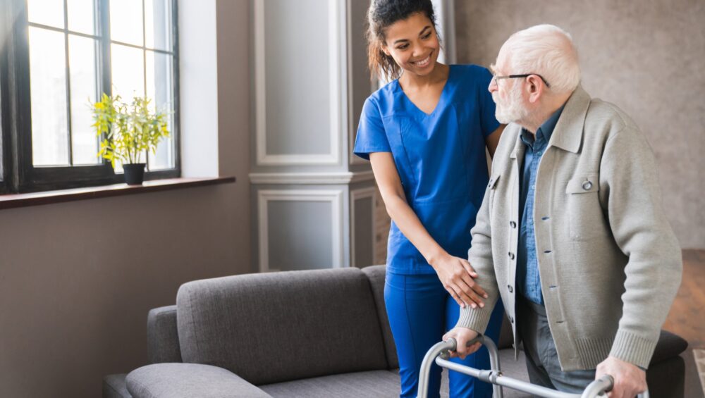 Caregiver in blue scrubs assisting elderly man using a walker inside a cozy living room with natural light.