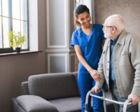 Caregiver in blue scrubs assisting elderly man using a walker inside a cozy living room with natural light.
