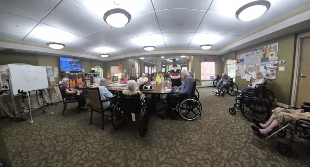 Elderly residents in wheelchairs gathered around a large table in a well-lit activity room of a care facility.
