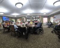 Elderly residents in wheelchairs gathered around a large table in a well-lit activity room of a care facility.