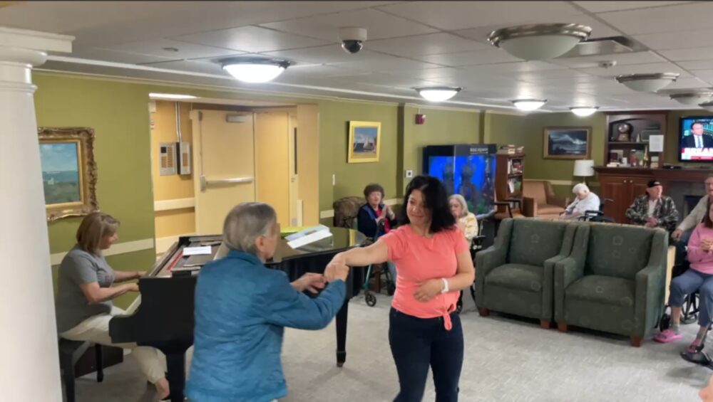 A female staff member and elderly female resident of Manoogian Manor dance in front of a woman playing the piano in the manor's living room.
