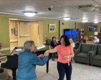 A female staff member and elderly female resident of Manoogian Manor dance in front of a woman playing the piano in the manor's living room.