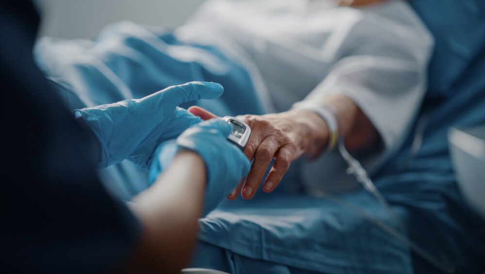 Healthcare worker wearing blue gloves placing a pulse oximeter on an elderly patient's finger in a hospital bed.
