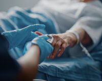 Healthcare worker wearing blue gloves placing a pulse oximeter on an elderly patient's finger in a hospital bed.