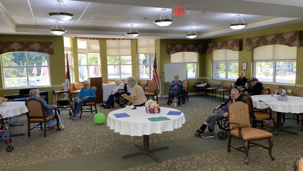 Residents at Manoogian Manor Home for the Armenian Aged playing indoor games. A group of elderly people seated in a spacious, well-lit community room with large windows and round tables covered with white cloths.