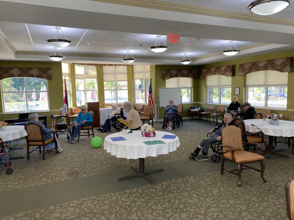 Residents at Manoogian Manor Home for the Armenian Aged playing indoor games. A group of elderly people seated in a spacious, well-lit community room with large windows and round tables covered with white cloths.