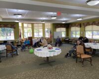 Residents at Manoogian Manor Home for the Armenian Aged playing indoor games. A group of elderly people seated in a spacious, well-lit community room with large windows and round tables covered with white cloths.