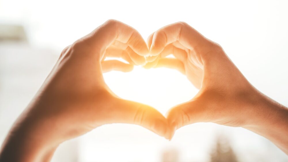 Two hands forming a heart shape with sunlight shining through the center against a blurred background.