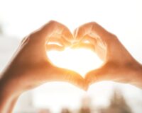Two hands forming a heart shape with sunlight shining through the center against a blurred background.