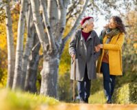 Elderly woman in gray coat and red beret walking with younger woman in yellow coat along a tree-lined path in autumn.
