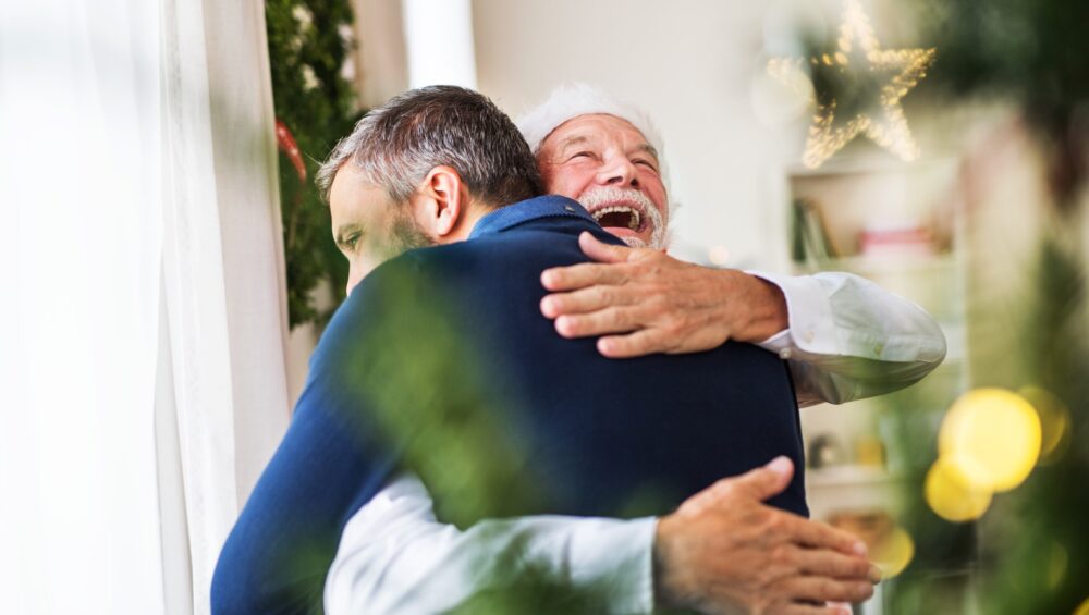A senior father with a Santa hat and adult son standing by the window at home at Christmas time, hugging.