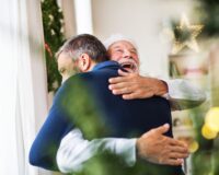 A senior father with a Santa hat and adult son standing by the window at home at Christmas time, hugging.
