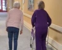 Two elderly women walking down a well-lit hallway at Manoogian Manor, one using a walker for support and the other walking independently.