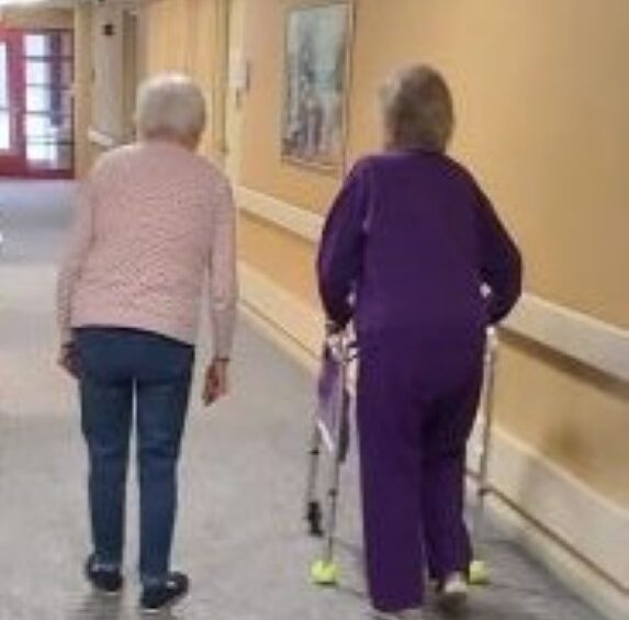Two elderly women walking down a well-lit hallway at Manoogian Manor, one using a walker for support and the other walking independently.