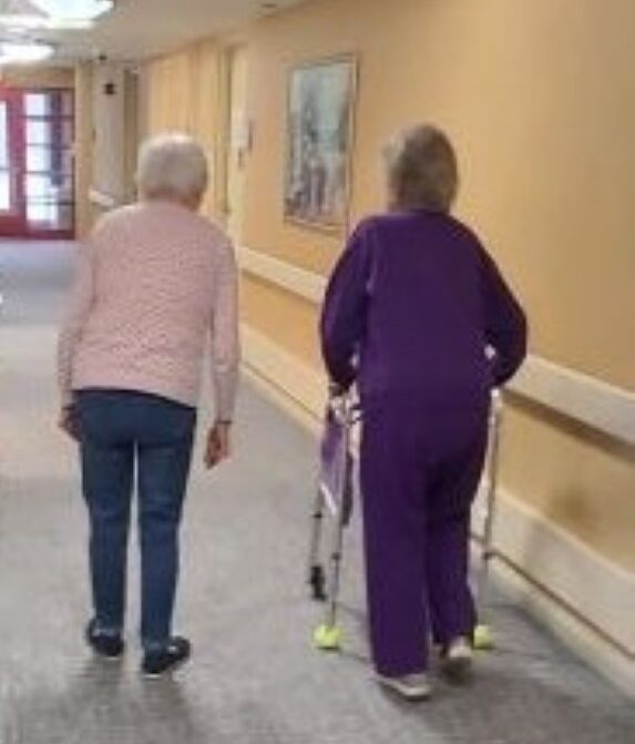 Two elderly women walking down a well-lit hallway at Manoogian Manor, one using a walker for support and the other walking independently.