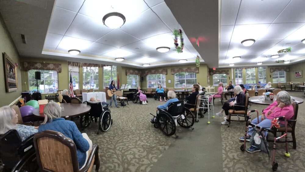 Elderly residents seated in a spacious common room at Manoogian Manor attentively listening to a man playing guitar and singing.