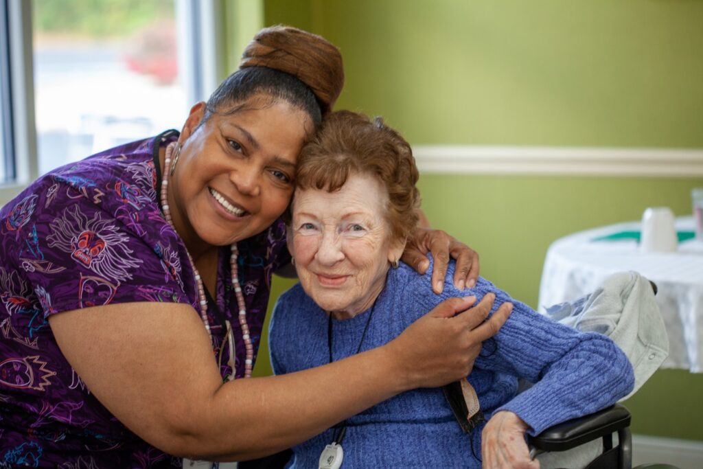 Caregiver in a purple patterned shirt warmly embraces an elderly woman in a blue sweater seated in a wheelchair inside a bright room.
