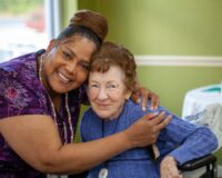 Caregiver in a purple patterned shirt warmly embraces an elderly woman in a blue sweater seated in a wheelchair inside a bright room.