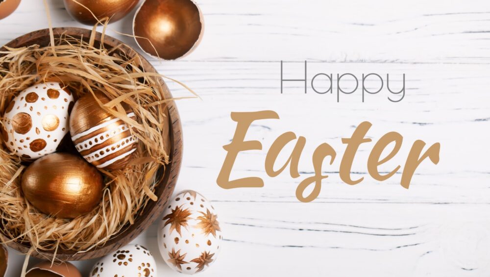 Decorative Easter eggs in gold and white with intricate patterns arranged in a wooden bowl filled with straw on a white wooden surface.