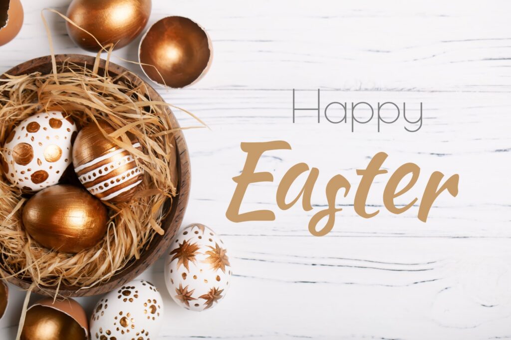 Decorative Easter eggs in gold and white with intricate patterns arranged in a wooden bowl filled with straw on a white wooden surface.