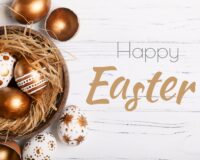 Decorative Easter eggs in gold and white with intricate patterns arranged in a wooden bowl filled with straw on a white wooden surface.
